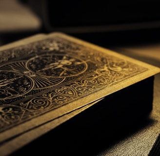 A deck of playing cards with intricate designs on the back, resting on a surface. The lighting is low and casts a warm, golden glow, highlighting the detailed patterns. The focus is on the textures and the beautiful craftsmanship of the card design.
