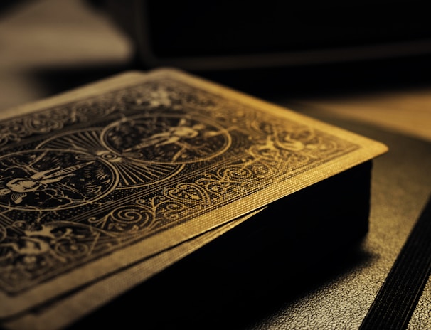 A deck of playing cards with intricate designs on the back, resting on a surface. The lighting is low and casts a warm, golden glow, highlighting the detailed patterns. The focus is on the textures and the beautiful craftsmanship of the card design.