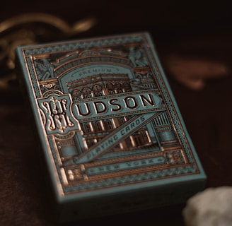 A decorative box of playing cards with intricate, vintage-style designs in gold and blue tones. The word 'Hudson' is prominently displayed on the front along with ornate patterns and architectural images. The background is dark, enhancing the elegance of the packaging.