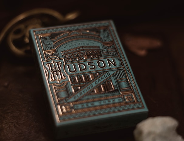 A decorative box of playing cards with intricate, vintage-style designs in gold and blue tones. The word 'Hudson' is prominently displayed on the front along with ornate patterns and architectural images. The background is dark, enhancing the elegance of the packaging.