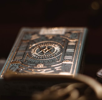 A deck of playing cards with intricate, metallic gold and teal designs featuring geometric patterns and a central emblem. The foreground is slightly out of focus, creating a bokeh effect, with other objects such as coins and a watch faintly visible in the background.