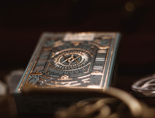 A deck of playing cards with intricate, metallic gold and teal designs featuring geometric patterns and a central emblem. The foreground is slightly out of focus, creating a bokeh effect, with other objects such as coins and a watch faintly visible in the background.
