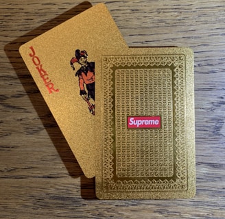 A pair of golden playing cards on a wooden table, one face displaying a joker with the word 'JOKER' printed vertically and the other showing an ornate pattern with a 'Supreme' logo in red.