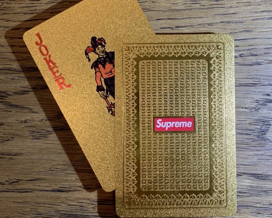 A pair of golden playing cards on a wooden table, one face displaying a joker with the word 'JOKER' printed vertically and the other showing an ornate pattern with a 'Supreme' logo in red.