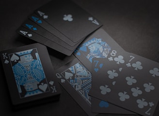 A set of playing cards with a dark and stylish design featuring black backgrounds and white and blue symbols. The cards include a King, Queen, and several numbered cards, all laid out on a dark surface.