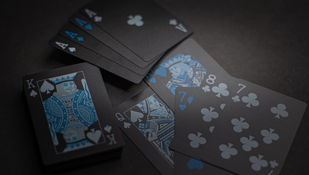 A set of playing cards with a dark and stylish design featuring black backgrounds and white and blue symbols. The cards include a King, Queen, and several numbered cards, all laid out on a dark surface.
