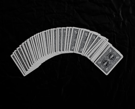 A neatly arranged spread of playing cards with an intricate design on their backs, set against a dark fabric background. The cards form a gentle curve, creating a sense of symmetry and order.