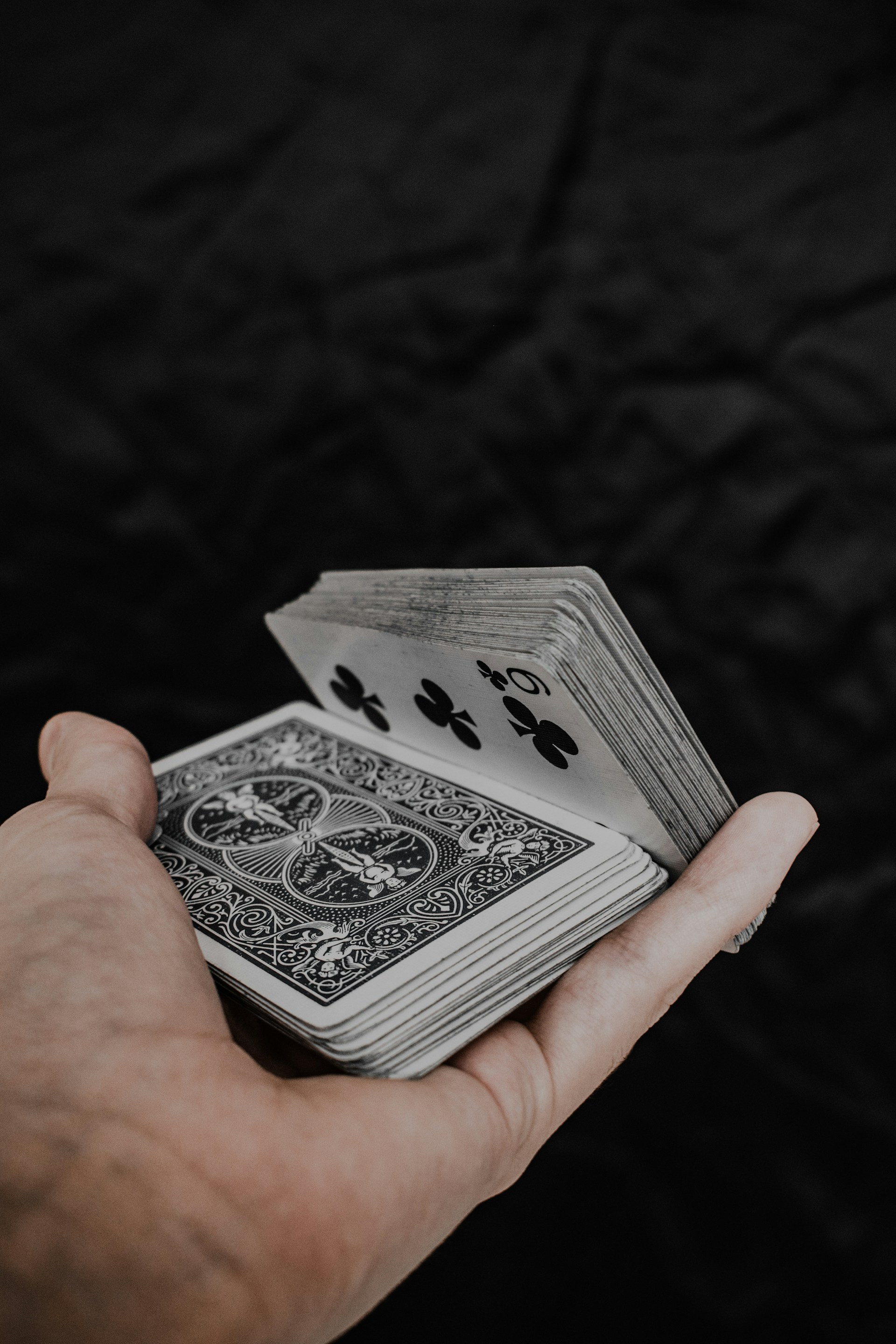 A hand holds a deck of playing cards with intricate back designs and a visible five of clubs. The background is a dark, textured surface, contrasting with the lighter tones of the cards and hand.