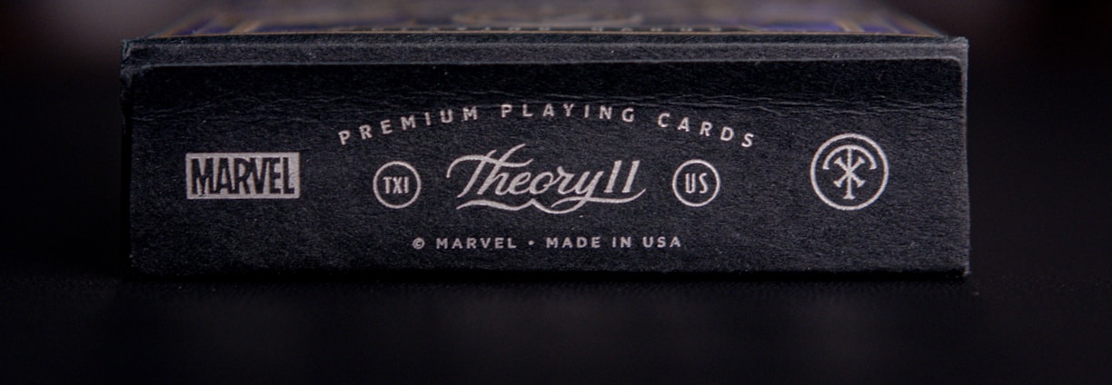 A close-up view of a black box of premium playing cards with the brand names Marvel and Theory11 prominently displayed. The design includes sleek typography and logo symbols, conveying a sense of luxury and exclusivity. The box is situated on a dark surface with a blurred background.