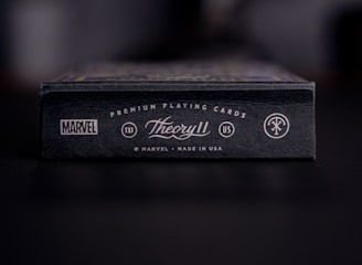 A close-up view of a black box of premium playing cards with the brand names Marvel and Theory11 prominently displayed. The design includes sleek typography and logo symbols, conveying a sense of luxury and exclusivity. The box is situated on a dark surface with a blurred background.