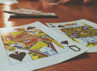 Playing cards are spread out on a wooden table, with the Queen and King of Spades prominently visible. The focus is on the vibrant colors and detailed designs of the cards, while other cards and hands are slightly blurred in the background, suggesting a game in progress.