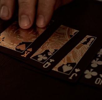 A hand gently resting on a spread of playing cards, featuring black suits and gold, ornate designs. The setting appears to be dimly lit, giving off a sophisticated and mysterious atmosphere.